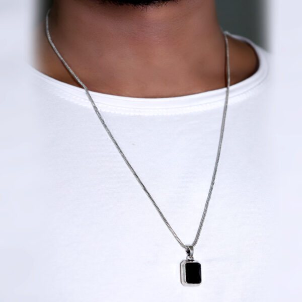 wearing a black stone pendant on a chain, visible against a white T-shirt. The sleek black stone contrasts with the white shirt, highlighting the pendant's elegant design and subtle sophistication."