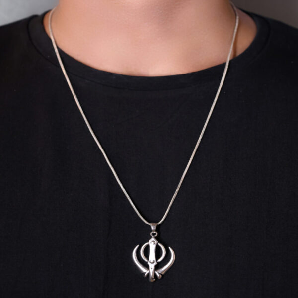 "Man wearing a Khanda pendant on a chain, visible against a black T-shirt. The pendant, featuring the Khanda symbol, stands out prominently against the dark background, emphasizing its detailed design."