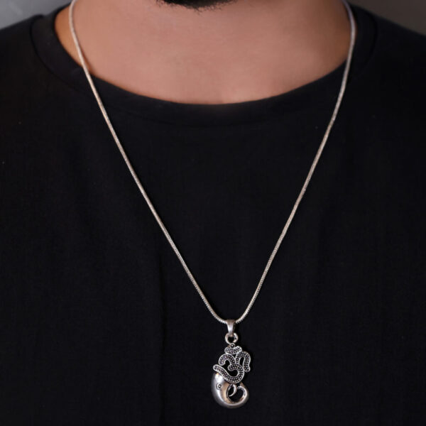 "Man wearing an Om Ganesha pendant with a chain, visible against a black T-shirt. The pendant, featuring a detailed Om symbol integrated with Ganesha's image, stands out prominently against the dark background, highlighting its spiritual significance."