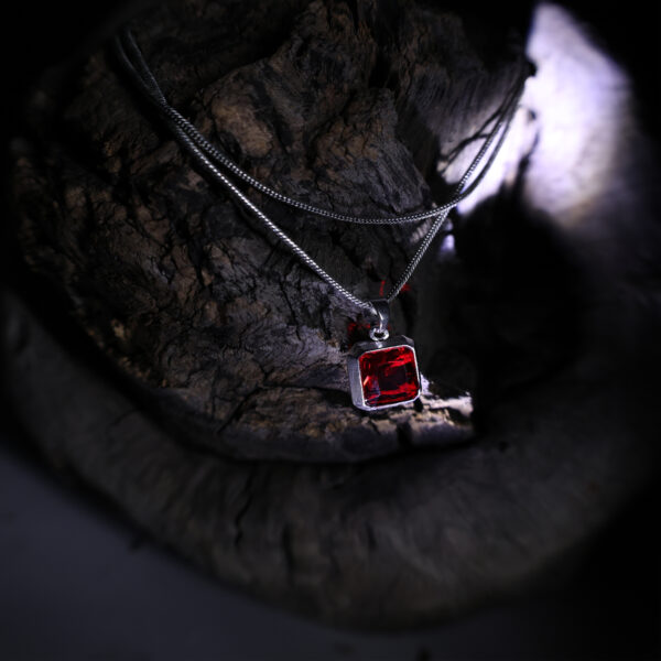 "Red stone pendant displayed on a dark wood background. The focus is on the red stone, which is illuminated to highlight its vibrant color and details against the textured, dark wood backdrop."