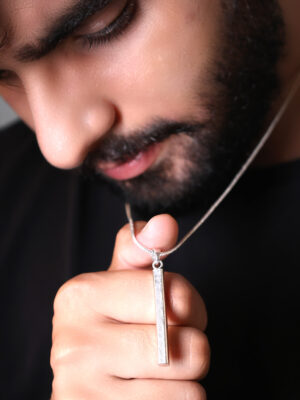 "Man wearing a silver thin bar pendant on a chain, visible against a black T-shirt. He is looking at the pendant, which is being focused by his thumb. The pendant is prominently displayed with the contrast between the silver and the black shirt."