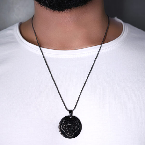 "Man wearing a black lion face pendant on a chain, visible against a white T-shirt. The pendant is prominently displayed in the center of the chest area of the shirt."