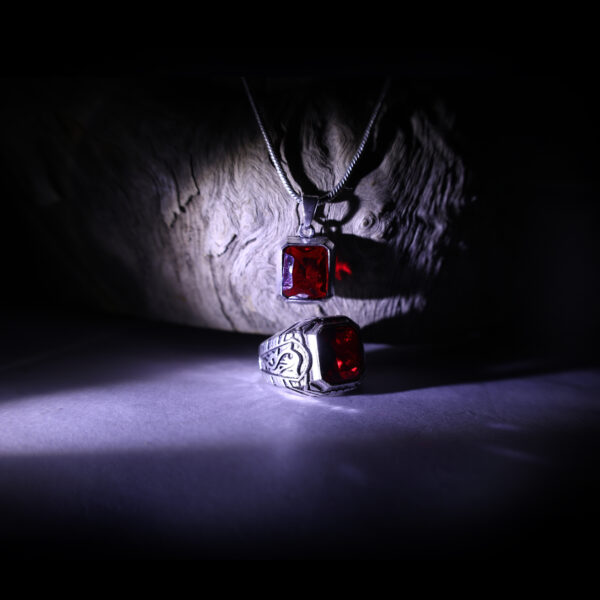 A red gemstone ring rests on a plain surface, while a pendant with a matching red gemstone hangs from a wooden surface, creating a contrasting display.