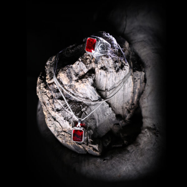 A silver-colored ring with a red gemstone and a matching pendant are displayed on a dark, textured wooden surface, illuminated by soft light with a moonlit background.