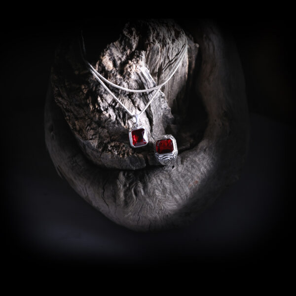 A silver-colored ring with a red gemstone and a matching pendant are displayed on a dark, textured wooden surface, illuminated by soft light with a moonlit background.