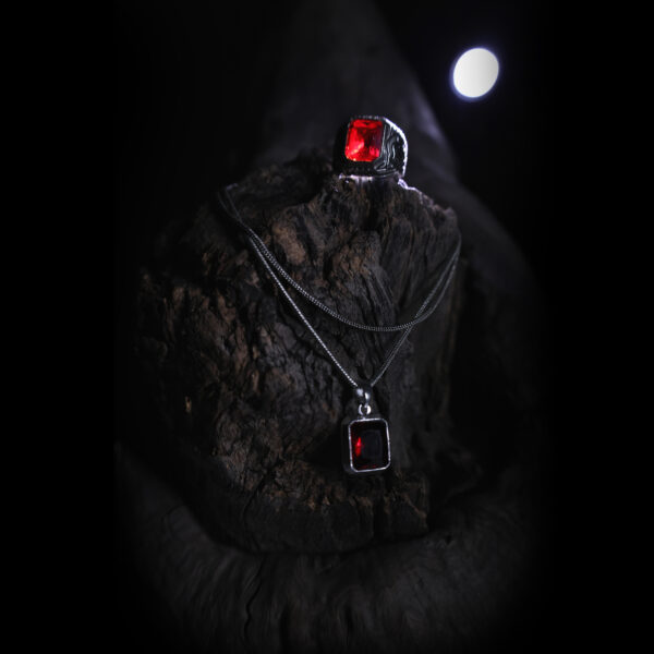 A silver-colored ring with a red gemstone and a matching pendant are displayed on a dark, textured wooden surface, illuminated by soft light with a moonlit background.