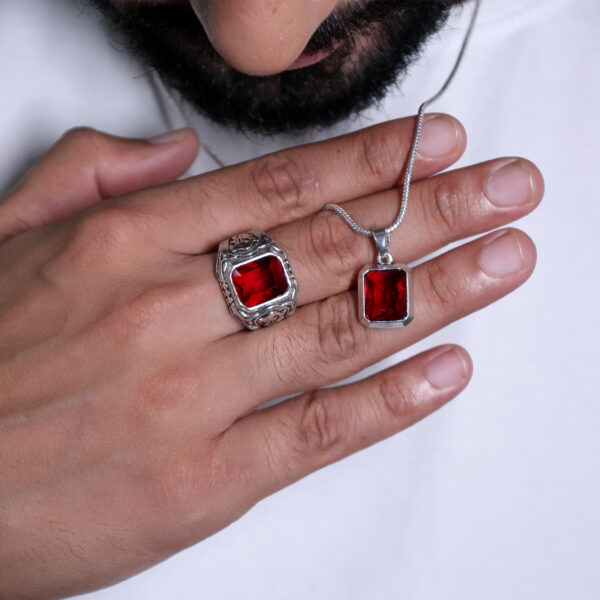 A close-up shot of a finger wearing a ring and pendant with large red gemstones, both featuring a silver-colored finish, set against a white background.