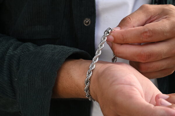 "A close-up of hands adjusting a braided silver-colored bracelet, worn on a wrist, with a green corduroy jacket and white t-shirt visible in the frame."